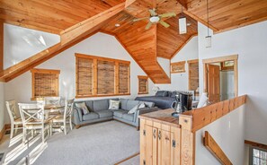 Une pièce de style « loft », avec un plafond haut, des poutres en bois, un canapé gris, une table à manger avec des chaises et un coin cuisine.