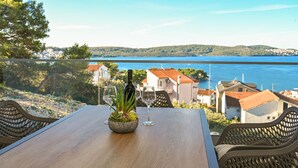 Une table en bois, ornée d’un pot de fleurs et de deux verres à vin, offre une vue sur une ville côtière, avec ses maisons et un ciel bleu dégagé.