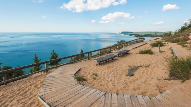 A wooden boardwalk leading to a sandy beach with benches and a railing overlooking a calm sea.