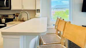 A kitchen with white cabinets, a stainless steel microwave, a sink, and a large window with a view of buildings and trees.