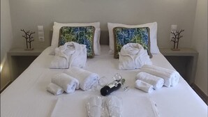 Cama bem arrumada com toalhas, um copo de água e uma garrafa, um quadro de uma árvore na parede e duas luminárias.