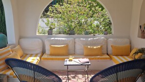 Eine sonnendurchflutete Terrasse mit einem weißen Sofa, gelben Kissen und zwei Korbstühlen. Vor dem Sofa steht ein kleiner Tisch mit einem Buch und einem Glas Orangensaft.