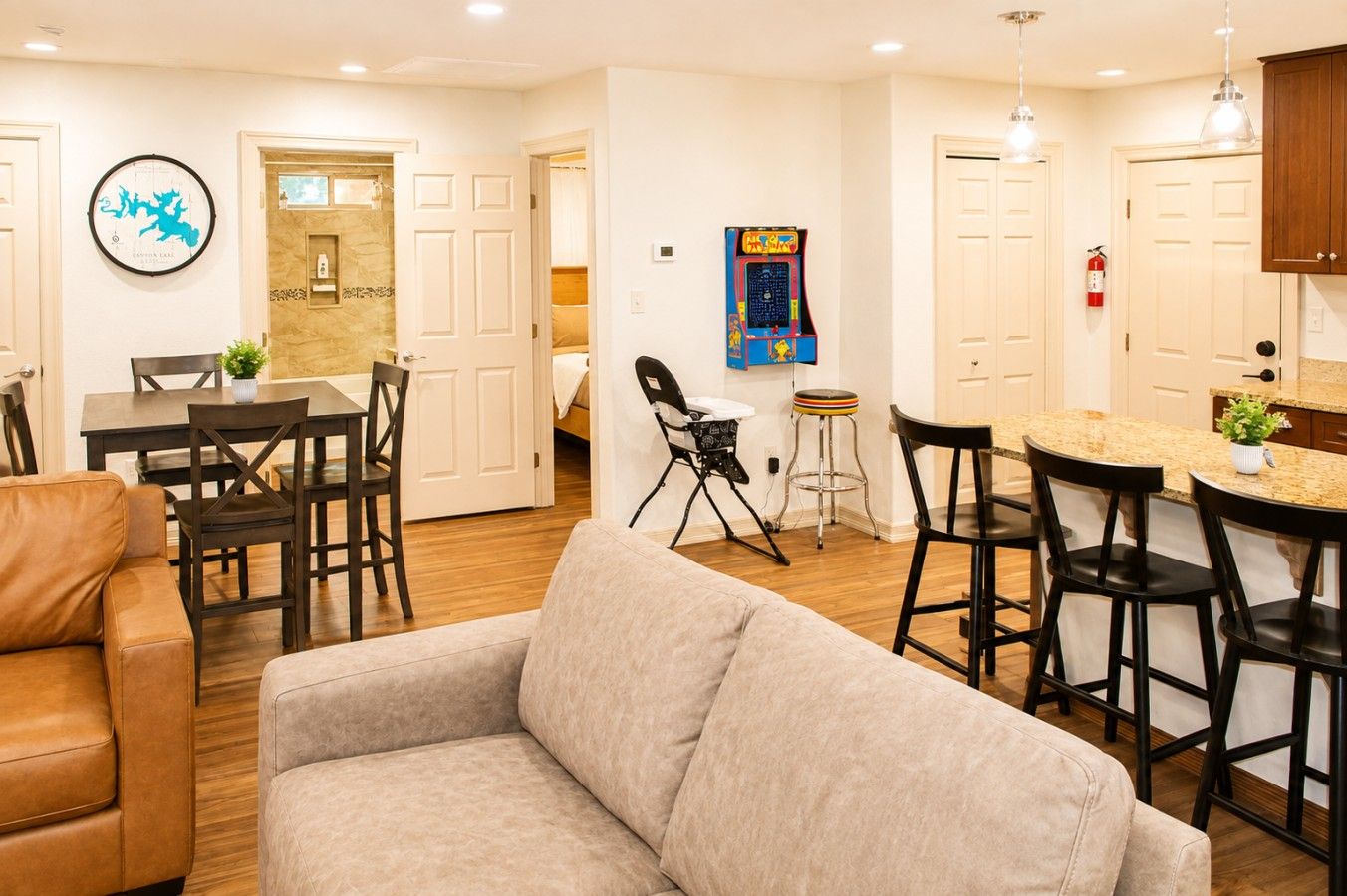 Un salon moderne avec un canapé, un coin repas avec des chaises, une cuisine avec des tabourets de bar et un jeu fixé au mur.