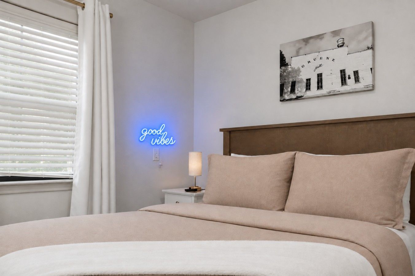 A bedroom with a bed, a bedside table, a lamp, a wall with a neon sign, and a picture on the wall.
