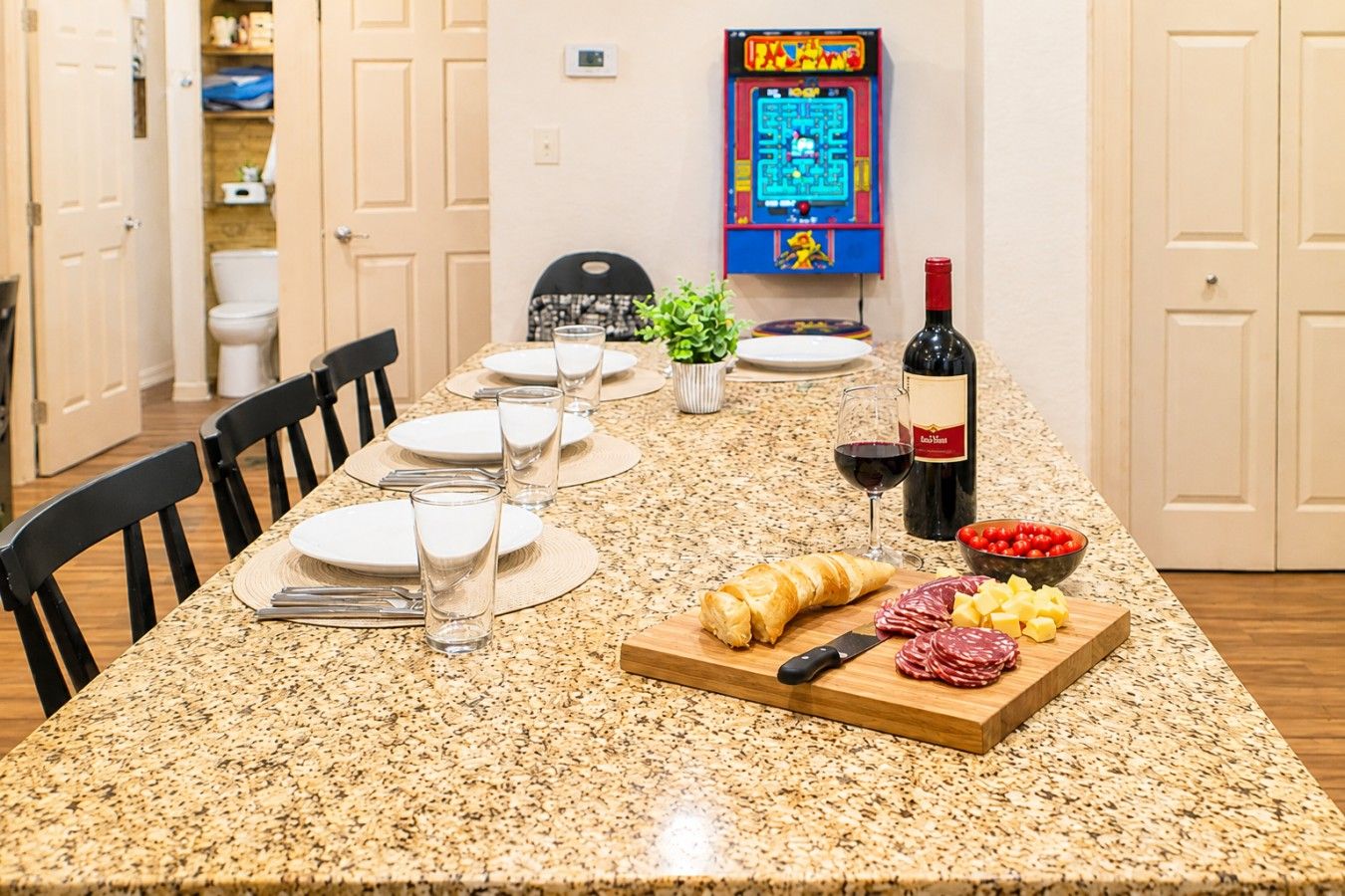 A dining table set with plates, glasses, and a bottle of wine, with a game machine in the background.