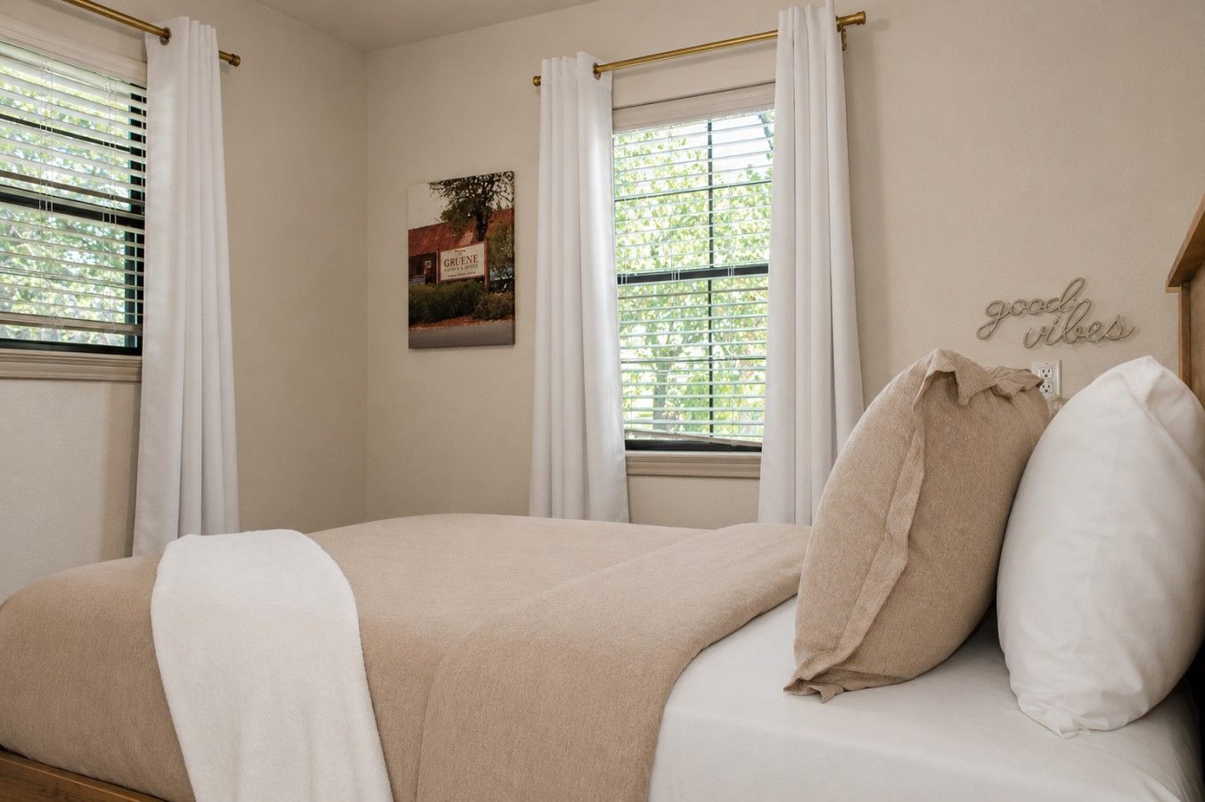 Une chambre à coucher avec un lit, des rideaux blancs, une fenêtre avec des stores, un panneau décoratif fixé au mur et un coussin décoratif.