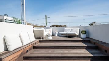 A rooftop terrace with a wooden deck, white outdoor furniture, and a potted plant.