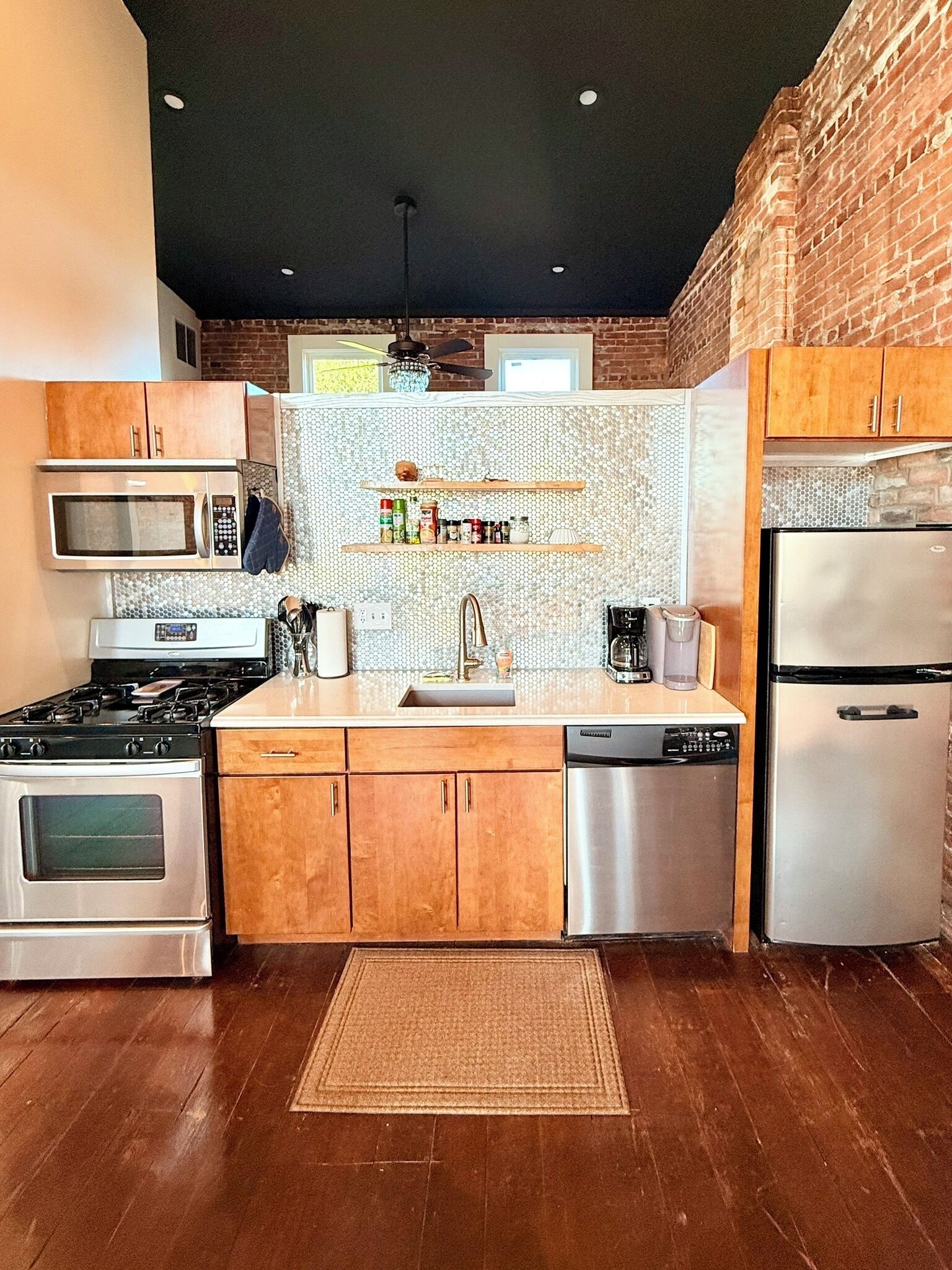 A modern kitchen with stainless steel appliances, wooden cabinets, a brick accent wall, and a tiled backsplash.