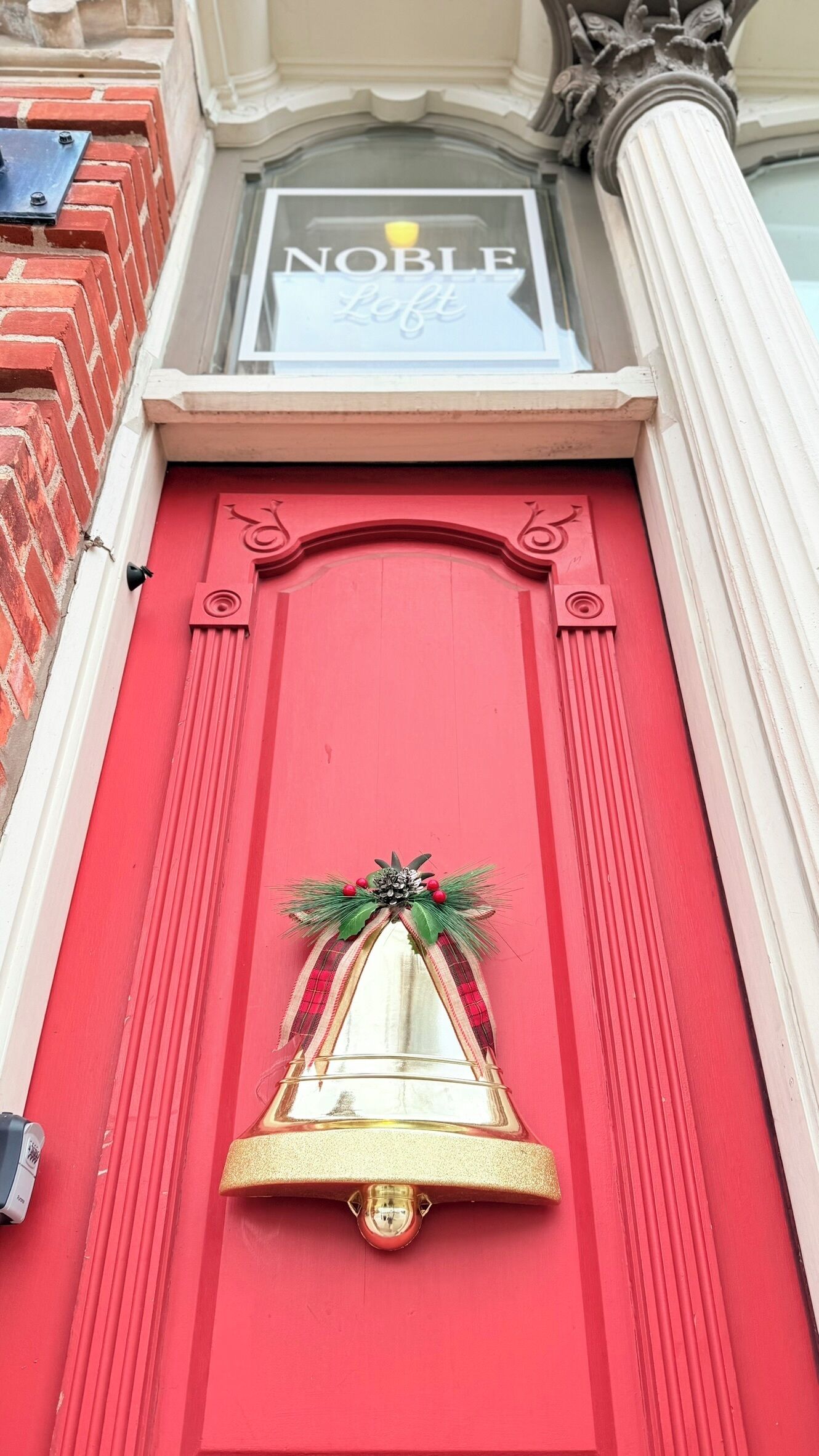 A red door with a golden bell and a wreath, above which is a sign reading "Noble Loft".