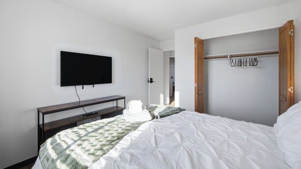 A hotel room with a bed, a flat-screen TV mounted on the wall, a wooden bench, and a coat rack with hangers. - Central Location B in Cul de Sac, 2 Bedroom, 1 Bath-sleeps 6, pets allowed (Kennewick)