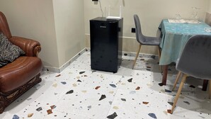 Une pièce avec un canapé en cuir marron, un réfrigérateur noir, une table recouverte d’une nappe bleue et un sol carrelé aux motifs multicolores.