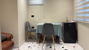 Un petit coin repas comprenant une table, des chaises, un réfrigérateur et une fenêtre avec des stores.