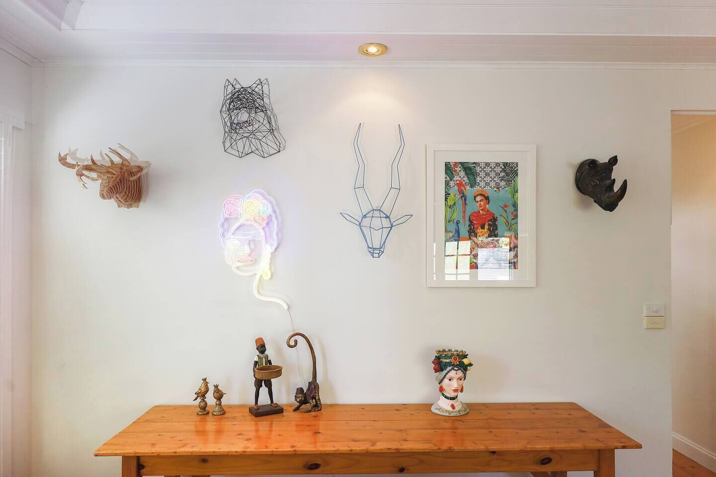 A room with a wooden table, a neon light, a framed painting, and various wall decorations including a skull and a geometric sculpture.