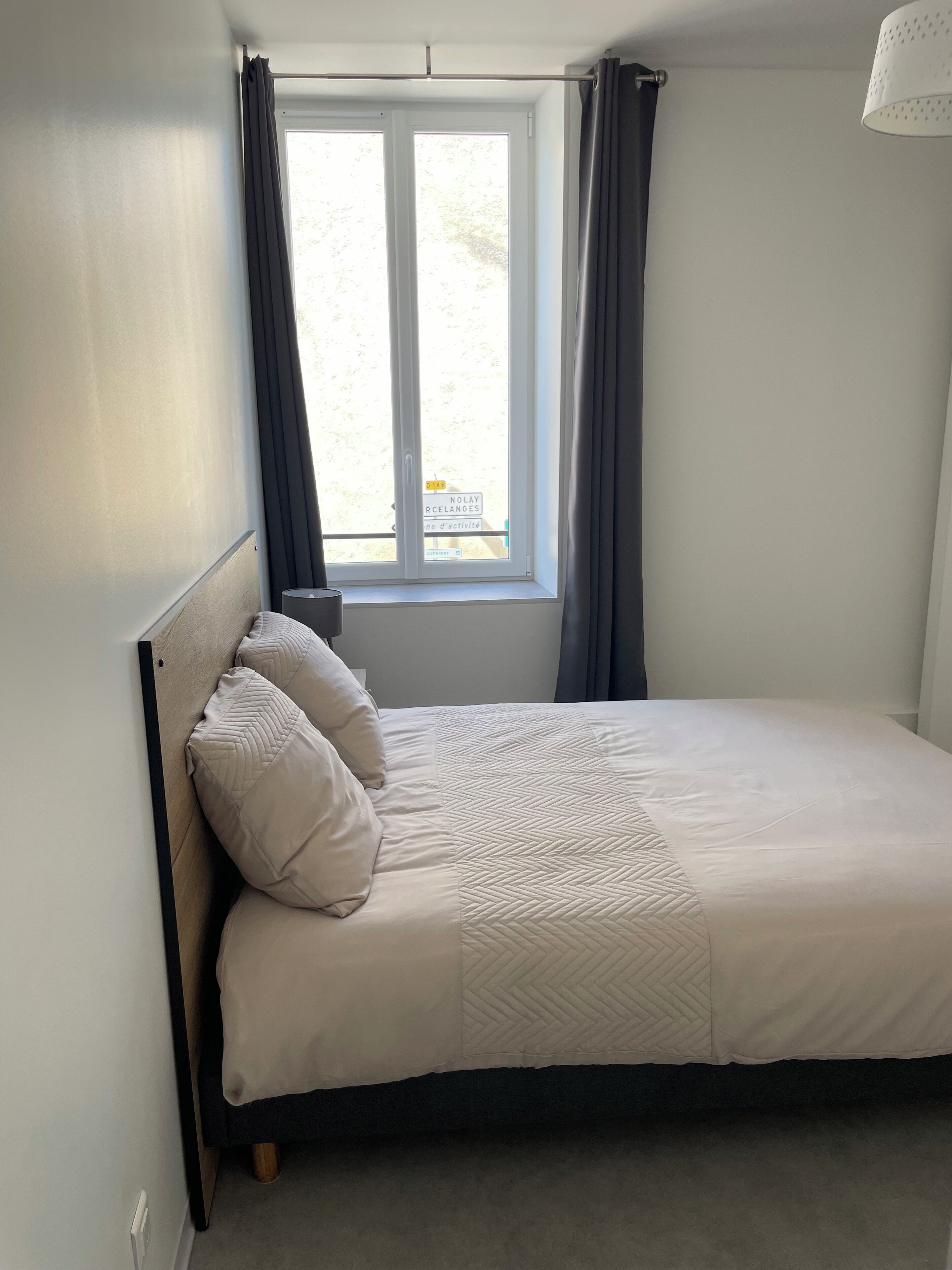 Un letto ben rifatto con una testiera, una finestra con tende e una lampada su un comodino.