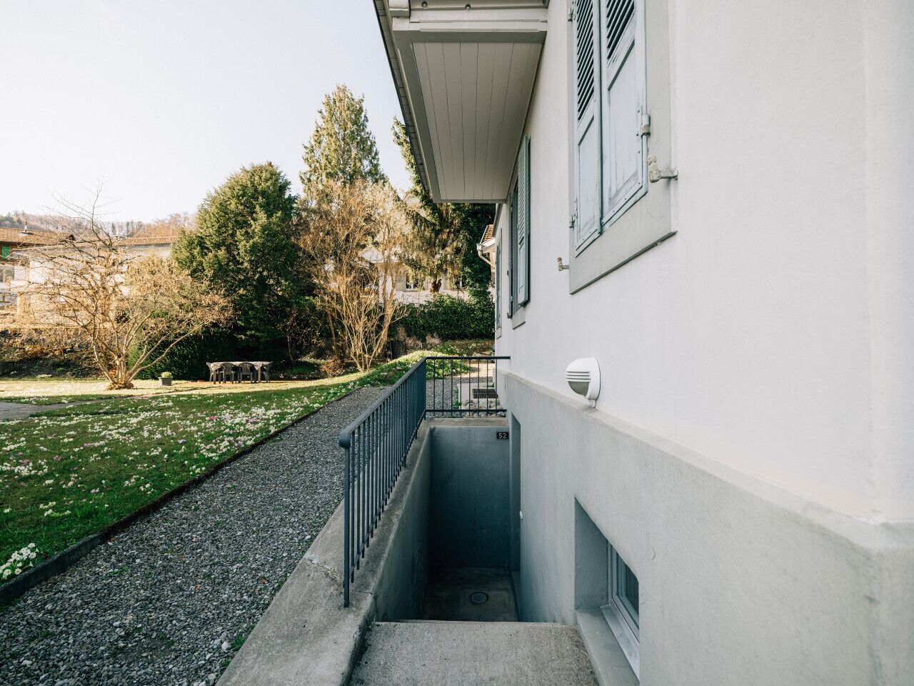 Un bâtiment doté d’un escalier extérieur étroit menant à une porte, le tout étant entouré d’une zone herbeuse et d’arbres.