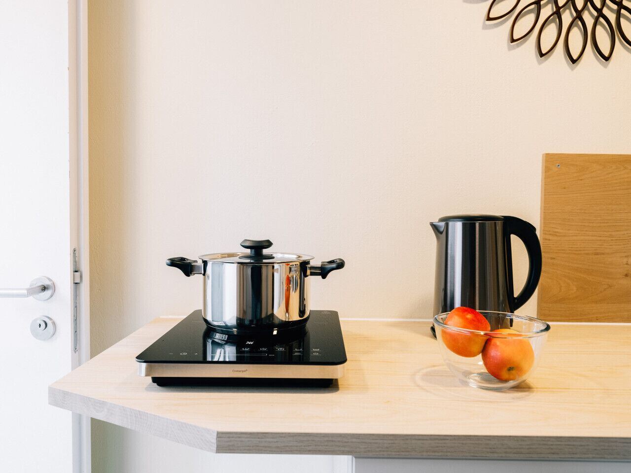 Una encimera de cocina con una cocina portátil de inducción, una olla de acero inoxidable, un bol de vidrio con manzanas y un hervidor eléctrico negro.