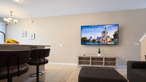 Una sala de estar moderna con un televisor de pantalla plana mostrando un paisaje urbano, una zona de bar con taburetes y una distribución de asientos acogedora.