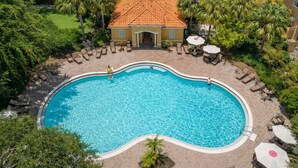 Una piscina con sillones de descanso y sombrillas, rodeada de palmeras y un edificio con tejado de baldosas.