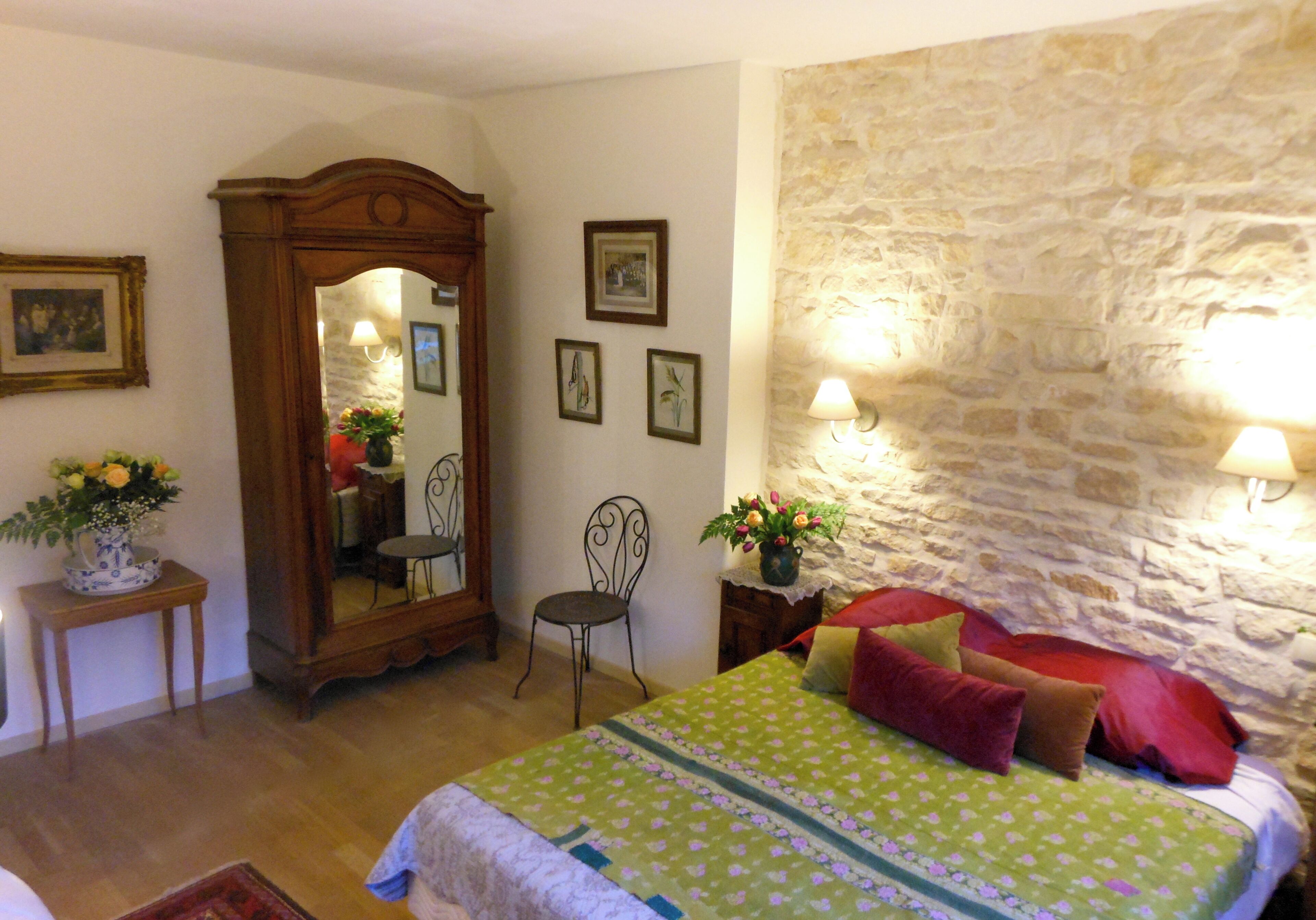 A bedroom with a stone wall, a bed with floral bedding, a wooden wardrobe, a mirror, and a small table with a vase of flowers.