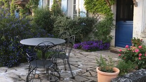 Een terras van natuursteen met een ronde tafel en stoelen, omringd door weelderige begroeiing en bloeiende planten.