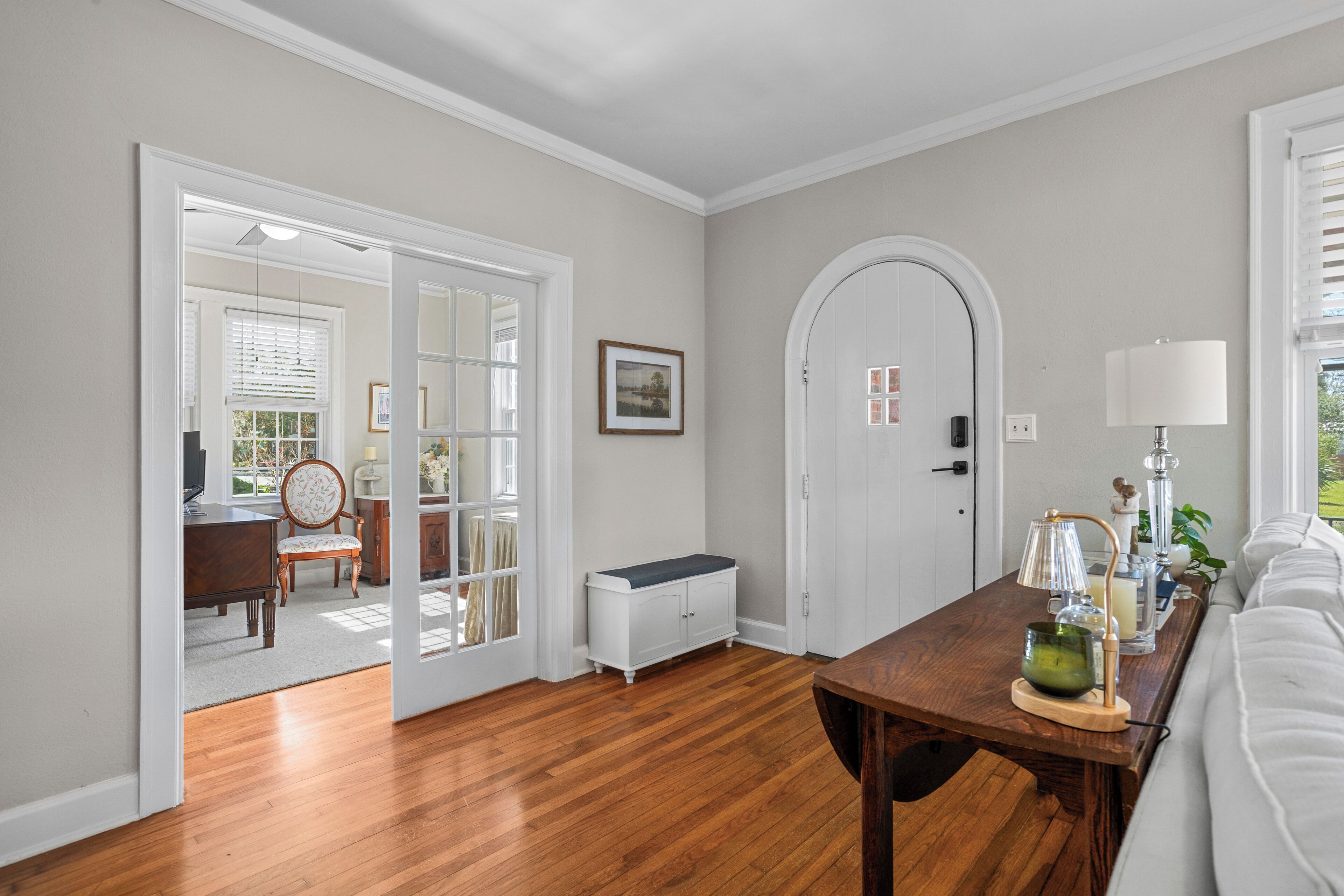 A room with a wooden floor, white walls, a white door, a bench, a table with a lamp, and a sofa.