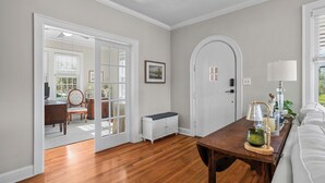 Una habitación con suelo de madera, paredes blancas, una puerta blanca, un banco, una mesa con una lámpara y un sofá.