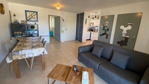 Een woonkamer met een grijze bank, een houten salontafel en een eethoek met een tafelkleed met patroon.