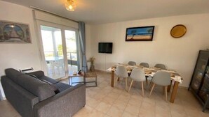 Een woonkamer met een bank, een eettafel met stoelen, een aan de muur bevestigde televisie en een schilderij.