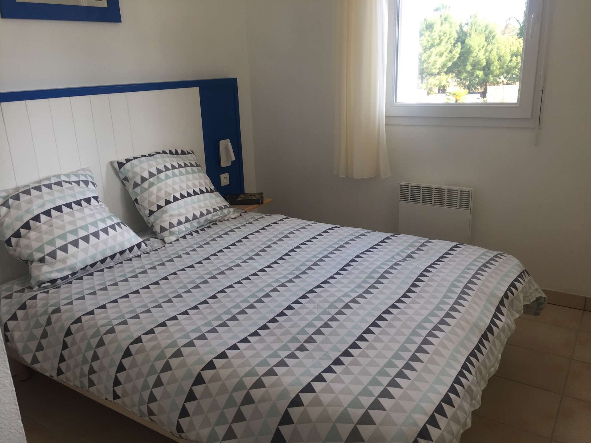 Ein Bett mit geometrisch gemustertem Überwurf, ein Nachttisch mit Lampe und ein Fenster mit Blick auf Bäume.