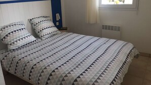 Een bed met een bedspreider met een geometrisch patroon, een nachtkastje met een lamp en een raam met uitzicht op bomen.