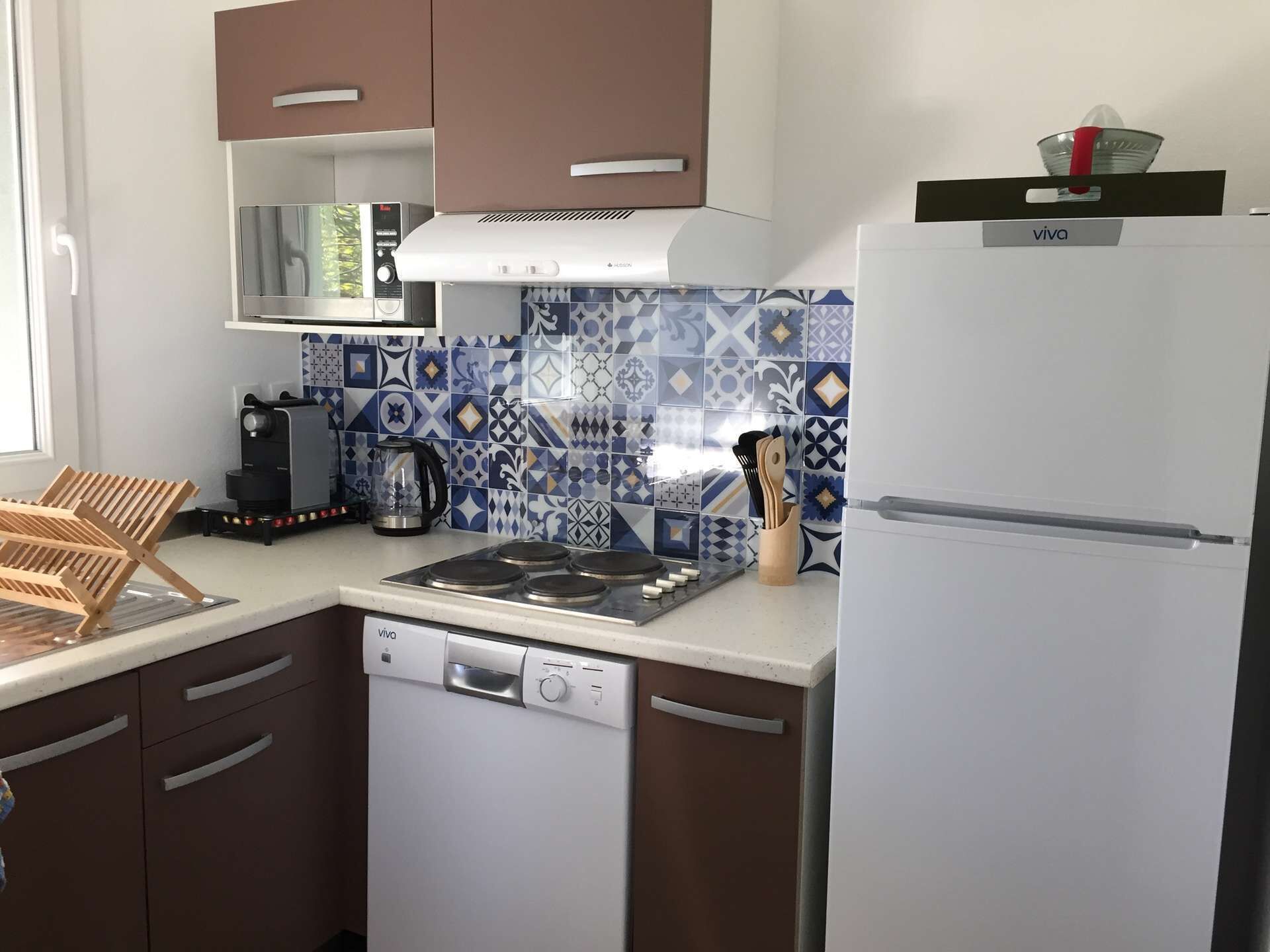 A compact kitchen with a white refrigerator, a stove, and a dishwasher. There is a coffee maker and a kettle on the counter. The kitchen has patterned tile backsplash and brown cabinets.