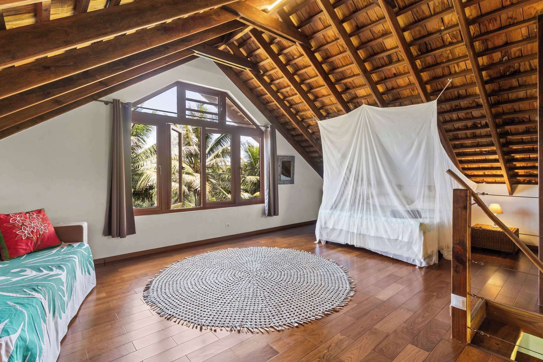 Una habitación con techo de madera, un ventanal con vistas a palmeras, una cama con almohada roja y un dosel blanco.