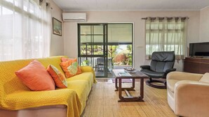 Ein Wohnzimmer mit gelber Couch, beigem Sessel, Couchtisch aus Holz und Fernseher. Eine Glastür führt zu einem Balkon mit Pflanzen und einem Tisch.