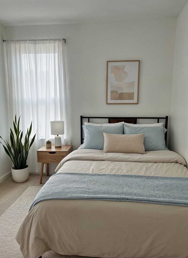 A bedroom with a bed, a bedside table with a lamp, a potted plant, and a framed picture on the wall.