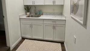 A small, white kitchen with a marble countertop, a sink, and cabinets. There is a rug on the floor and a framed picture on the wall. - Austin Living at Picnic Dr , Modern, Private & Bright (Round Rock)