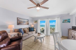 A living room with a sofa, a coffee table, a white chair, a ceiling fan, and a view of the ocean.