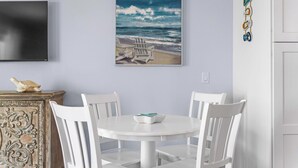 Een eetruimte met een ronde witte tafel en vier witte stoelen, een schilderij van een strand en een decoratieve wandhanger.