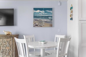 Ein Essbereich mit einem runden weißen Tisch, vier weißen Stühlen, einem Strandbild und einem Wandbehang.