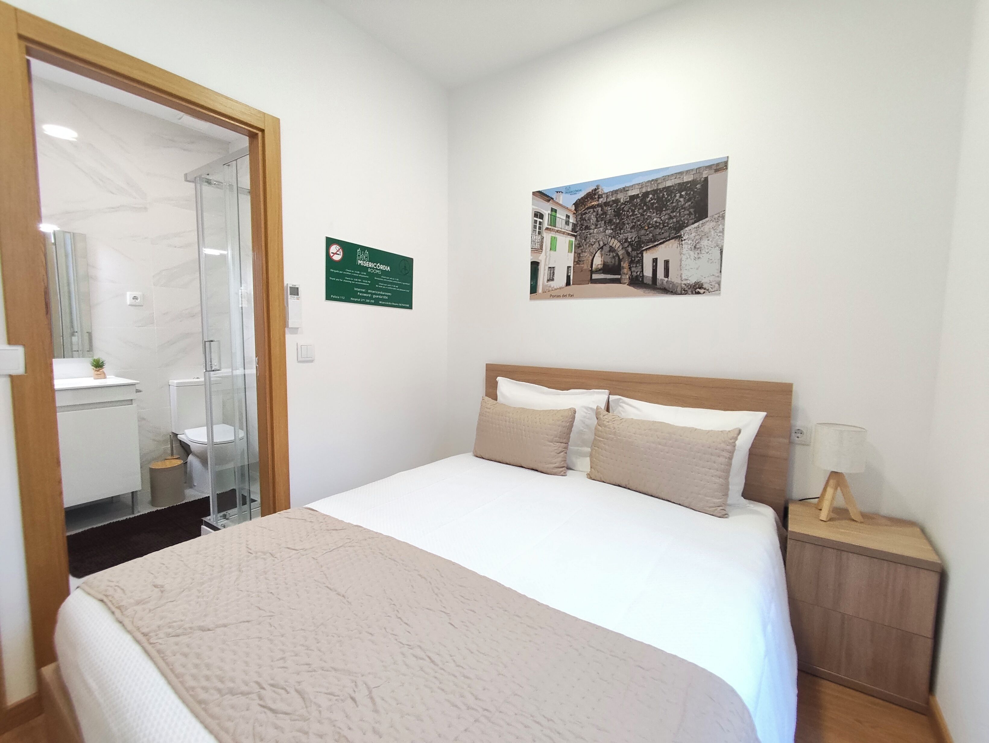 Un dormitorio con cama, mesita de noche, un cuadro en la pared y un baño visible a través de una puerta abierta.