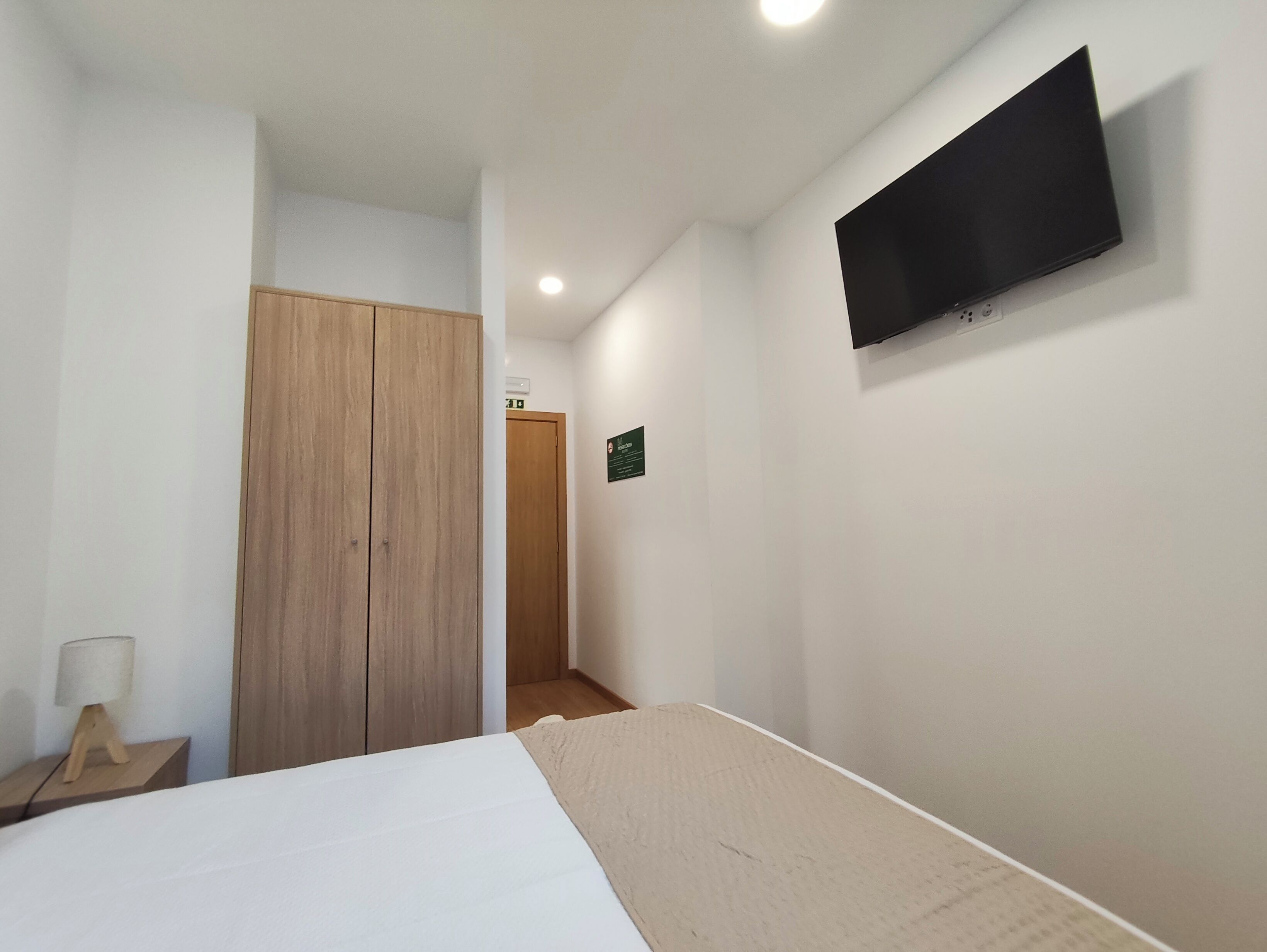 Une chambre à coucher comprenant un lit, une armoire en bois, un téléviseur fixé au mur et une petite table de chevet avec une lampe.