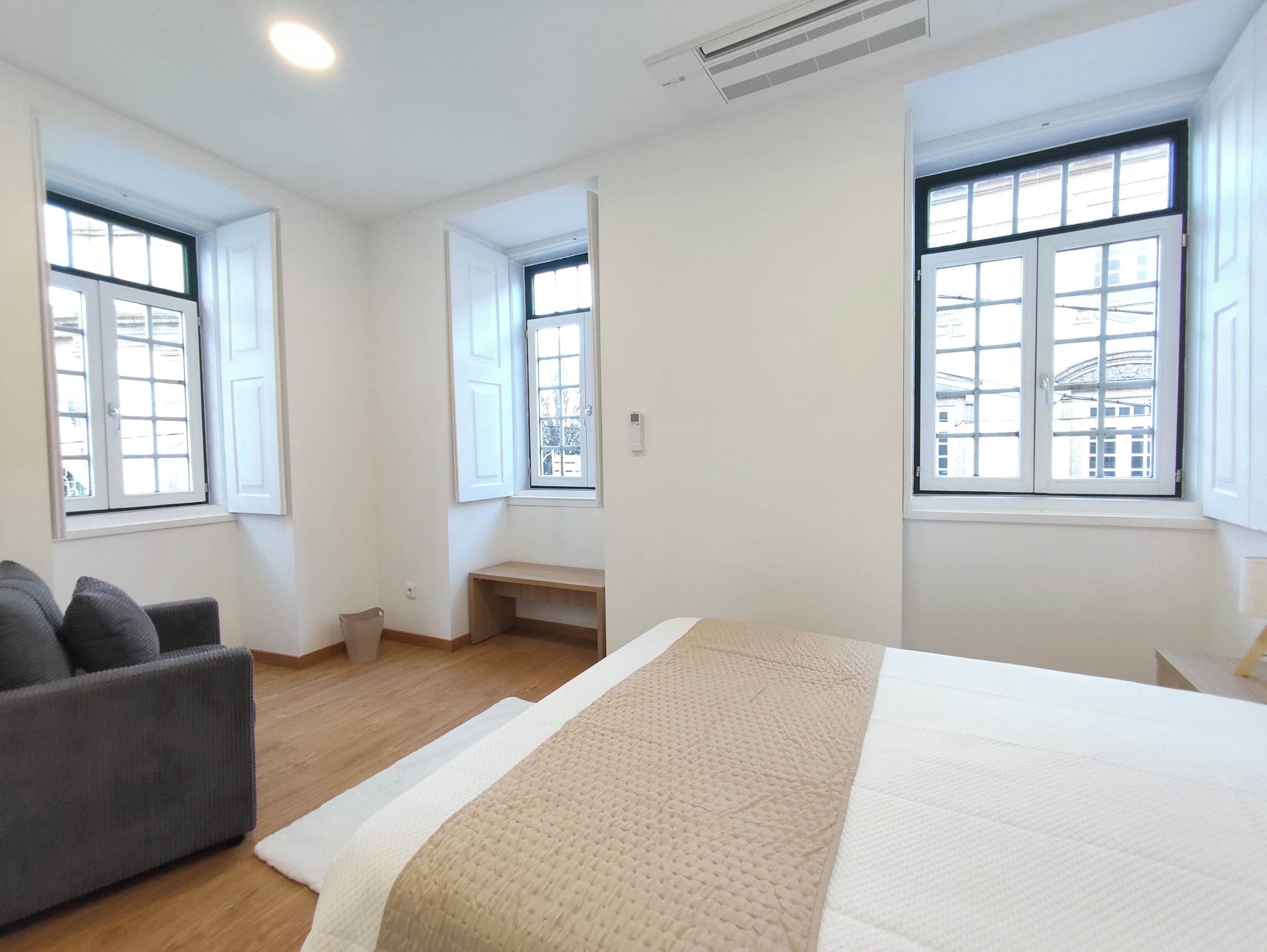 Ein Schlafzimmer mit einem Bett, einem grauen Sofa und zwei Fenstern mit Blick auf Gebäude.