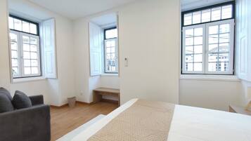 Ein Schlafzimmer mit einem Bett, einem grauen Sofa und zwei Fenstern mit Blick auf Gebäude.