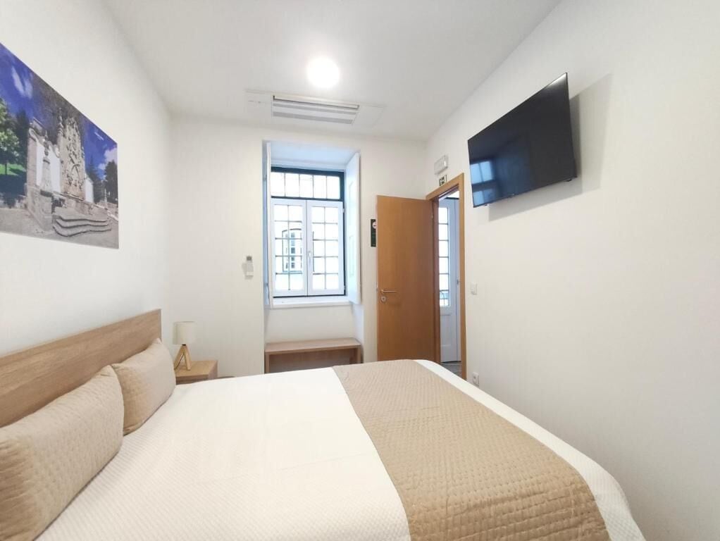 Habitación de hotel con una cama, televisor montado en la pared, ventana y puerta que da a otra habitación.