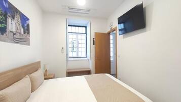 Habitación de hotel con una cama, televisor montado en la pared, ventana y puerta que da a otra habitación.