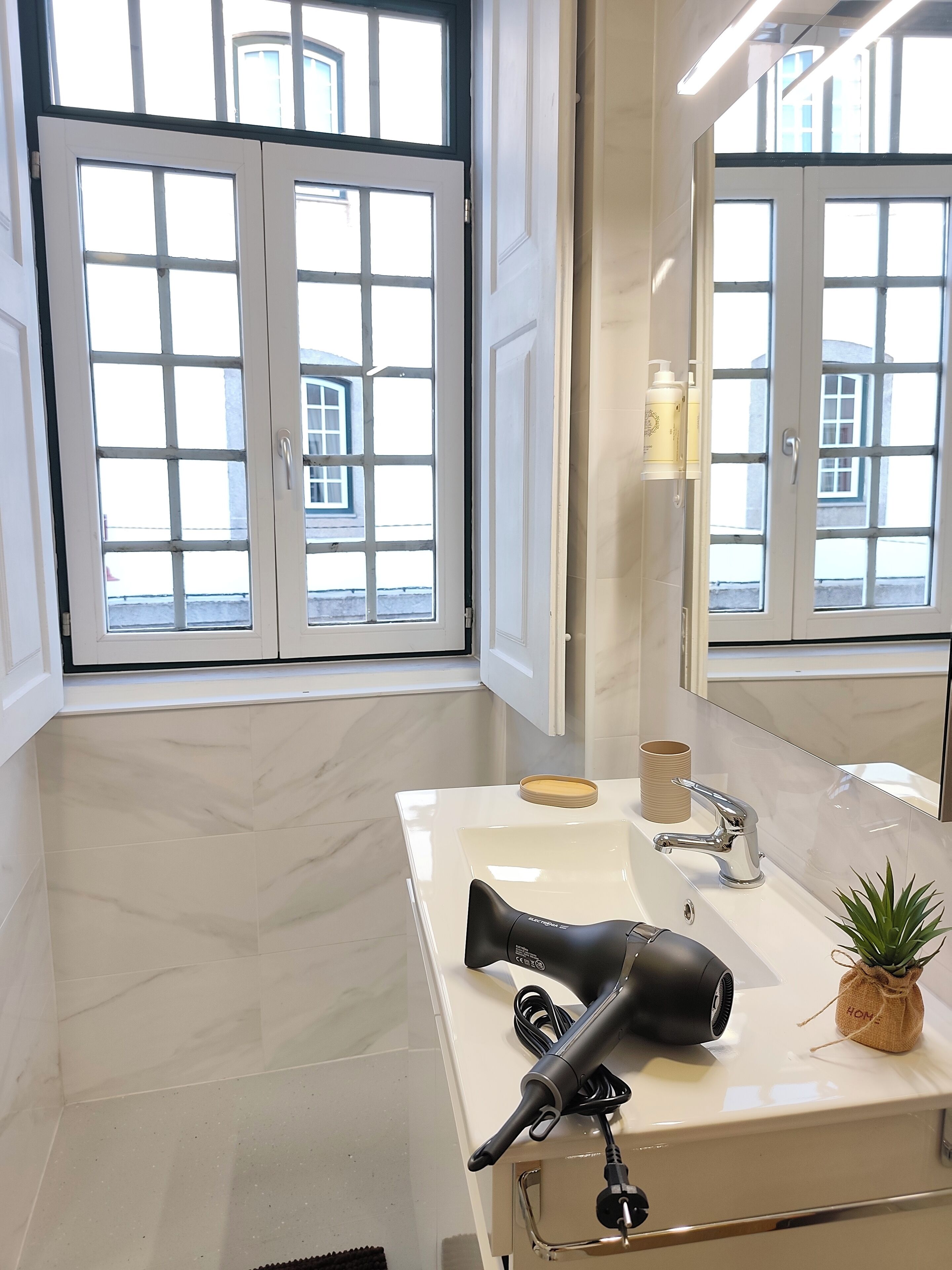 Um banheiro com piso de mármore branco, uma pia branca com torneira moderna, um secador de cabelo e um pequeno vaso de planta.
