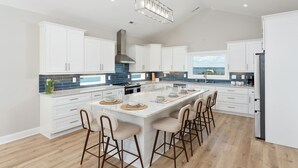 Une cuisine moderne avec des meubles blancs, un grand îlot central doté de tabourets, et une vue sur la mer par la fenêtre.