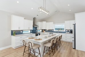 A modern kitchen with white cabinets, a large island with bar stools, and a view of the sea through the window. - Luxury Beach Retreat, 7BR, Dock, 360° Views (North Topsail Beach)