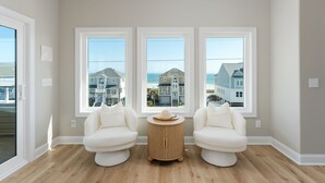 Une pièce dotée de trois grandes fenêtres, d’une table ronde en bois et de deux fauteuils blancs.