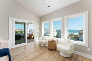 A spacious living room with large windows, a wooden floor, and a view of the ocean and buildings. - Luxury Beach Retreat, 7BR, Dock, 360° Views (North Topsail Beach)