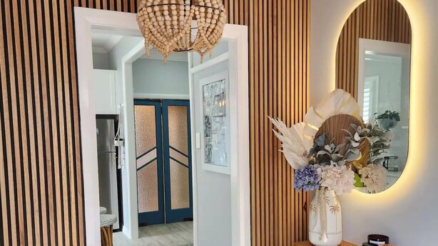 A hallway with vertical wooden slats, a woven light fixture, a mirror with a circular frame, and a vase with flowers on a blue cabinet.